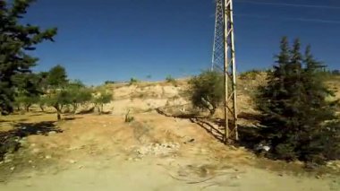 Hareket eden bir arabanın açık mavi gökyüzünün altında kurumuş ve kayalık bir araziyi çevreleyen yeşil ağaçlarla dolu manzaralı bir manzara. Suriye 'de.