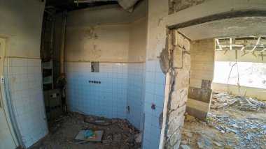 Dökülen duvarları ve kırılan fayansları olan terk edilmiş bir oda harap bir iç mekanı ortaya çıkarıyor. Zemin molozlarla doludur ve güneş ışığı bir açıklıktan akar, ihmalkarlığın altını çizer..