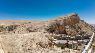 Açık mavi bir gökyüzünün altında kayalık bir uçuruma kurulmuş küçük bir köyün olduğu panoramik bir manzara. Maaloula, Suriye.
