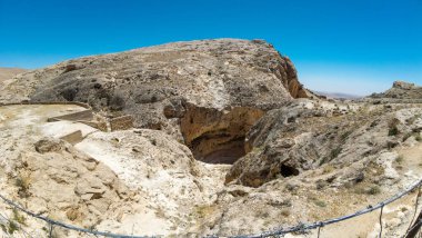 Kurak arazi ve açık mavi gökyüzü ile çevrili büyük bir mağara girişinin yer aldığı kayalık bir manzaranın panoramik görüntüsü. Sahne, doğal çevrenin engebeli güzelliğini yakalar..