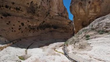 Dar, sarp duvarları ve açık mavi gökyüzünün altında kurumuş bir nehir yatağı olan kayalık bir kanyon..