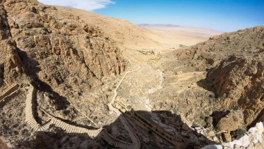 Kayalık bir çöl manzarası, dolambaçlı yolları ve berrak mavi gökyüzünün altındaki uzak dağları gösteriyor..