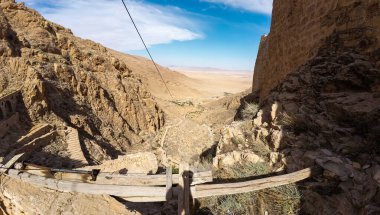 Açık mavi gökyüzünün altında kayalık uçurumlar ve antik kalıntılarla dolu bir çöl manzarası..