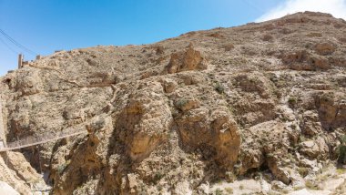 Kayalık tepeleri ve kanyonu geçen dar asma köprüsü olan engebeli bir çöl manzarası..