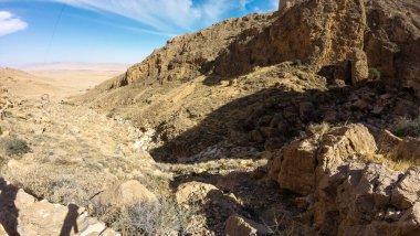 Mavi gökyüzünün altında kayalık uçurumlar ve seyrek bitki örtüsü olan engebeli bir çöl manzarası..