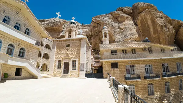 Kayalık bir uçurumun kenarına kurulmuş tarihi bir manastır manzarası. Üzerinde taş mimarisi ve çatılarda açık mavi bir gökyüzünün altında haçlar var..