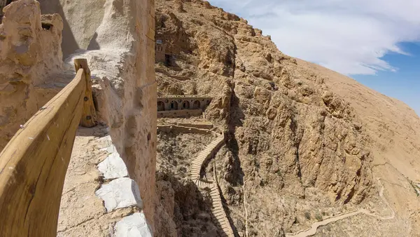 Engebeli bir yamaç boyunca esen ahşap korkuluklu dar bir taş yol kurak manzarayı ve kayaların içine inşa edilmiş antik yapıları çarpıcı bir şekilde gösteriyor..