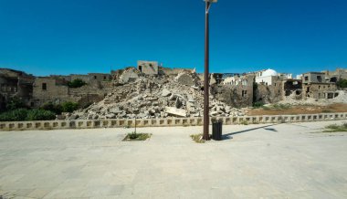 Açık mavi gökyüzünün altında yıkılmış binalar, önünde enkaz ve sokak lambası var. Halep, Suriye