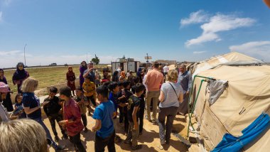 22/05/2025. Halep, Suriye yakınlarındaki mülteci kampı. Aralarında çocuklar ve yetişkinlerin de bulunduğu bir grup insan, açık bir gökyüzünün altındaki çadırların yakınında toplanıyor. Bir grup mülteci ziyaretçi yardımcılarla buluşuyor.