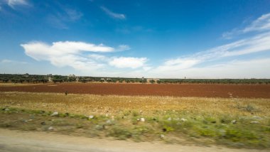 Açık mavi gökyüzü, dağınık bulutlar ve kırmızı topraklardan oluşan geniş bir kırsal alan. Kırsal Suriye