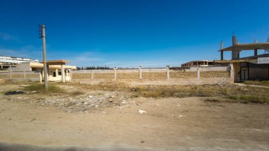 Kısmen inşa edilmiş bir bina ve açık mavi gökyüzünün altında uzun tuğla bir duvar olan çorak bir arazi. Kırsal Suriye