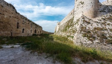 Mavi gökyüzünün altında yüksek duvarları ve çimenli hendekleri olan tarihi bir taş kale. Krak des Chevaliers kalesi, Suriye