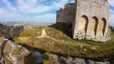 Tepenin tepesinde kemerli pencereleri olan, çimenli bir manzara ve açık mavi gökyüzüyle çevrili antik taş kale. Krak des Chevaliers kalesi, Suriye