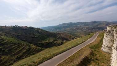 Parçalı bulutlu bir gökyüzünün altındaki yuvarlanan tepelerin arasından dolambaçlı bir yolun manzarası. Homs Boşluğu, Suriye