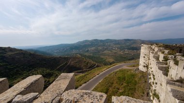 Yuvarlanan tepelere ve vadilere bakan tarihi bir kaleden panoramik bir manzara. Manzara, parçalı bulutlu bir gökyüzünün altında dolambaçlı yollar ve uzak dağlara sahiptir. Homs Boşluğu, Suriye