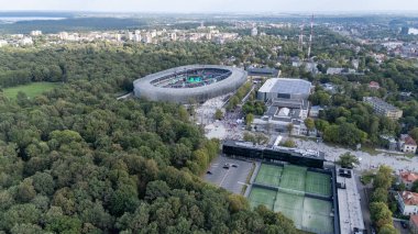 Yeşilliklerle çevrili, arka planda kentsel binalarla çevrili modern bir stadyumun havadan görünüşü. Sahne, stadyumun dışında toplanan insanlarla canlı bir atmosfer yakalıyor..