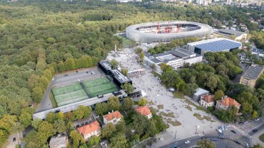 Geniş bir stadyum, tenis kortları ve yakınlardaki binaların bulunduğu yemyeşil alanlarla çevrili bir spor kompleksinin havadan görünüşü. İnsanlar açık meydanda toplanmış, bir etkinlik öneriyorlar..