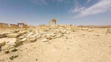 IŞİD tarafından mavi gökyüzünün altında bulutlu bir çöl manzarasında yıkılmış antik kalıntılar. Suriye, Palmira.