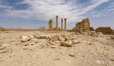 IŞİD tarafından mavi gökyüzünün altında bulutlu bir çöl manzarasında yıkılmış antik kalıntılar. Suriye, Palmira.