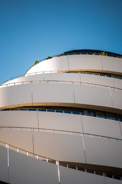 Çarpıcı bir modern bina kıvrımlı, katmanlı beyaz bir cephe ile çerçeveye hükmediyor, açık mavi gökyüzüne karşı yükseliyor.