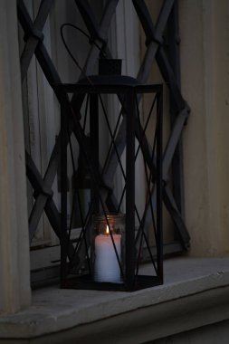 İçinde parlayan bir mum olan siyah metal bir fener pencere pervazında ya da çıkıntıda durur. Alevin sıcacık ışığı çevreyi aydınlatır.