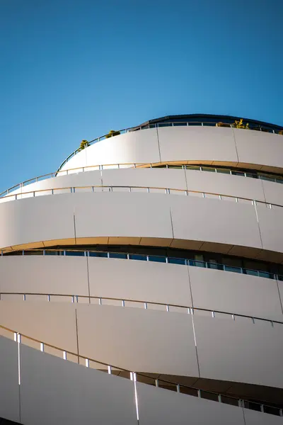 Çarpıcı bir modern bina kıvrımlı, katmanlı beyaz bir cephe ile çerçeveye hükmediyor, açık mavi gökyüzüne karşı yükseliyor.