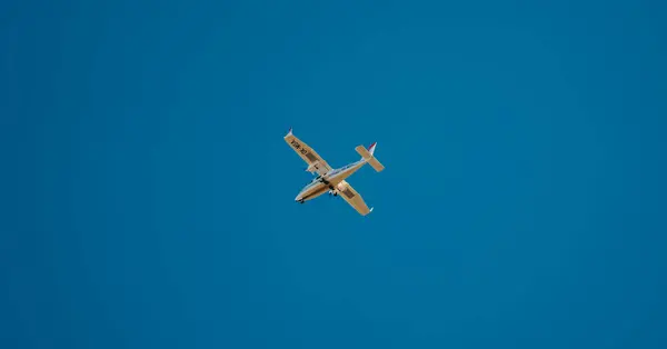 Küçük, açık renkli bir uçak engin ve berrak bir mavi gökyüzünde uçuyor, aşağıdan görülüyor. Kuyruğunda ince kırmızı aksanlı beyaz ya da açık gri bir uçak.