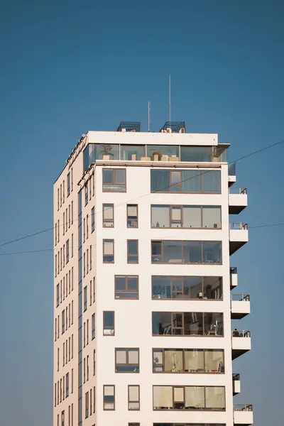 Çok sayıda dikdörtgen penceresi ve balkonları olan uzun, modern bir beyaz bina, açık mavi gökyüzüne karşı alçak bir açıdan görülüyor..
