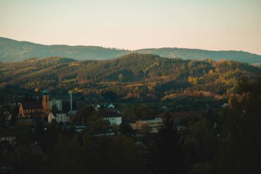 Geniş bir panoramik manzara, sakin bir vadi boyunca uzanır, geniş, ormanlı dağların eteklerinin arasında kısmen gizlenmiş bir kasabayı ortaya çıkarır. Gökyüzü, ufuktaki sıcak, ruhani bir portakaldan daha yumuşak, daha hafif bir tona dönüşüyor. 