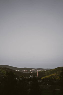 Üst çerçeveye hakim soluk, bulutlu bir gökyüzünün geniş genişliği, aşağıdaki manzaranın üzerine yumuşak ve dağınık bir ışık yayar. Midground 'da, bir vadide yuva yapmış uzak bir şehir görülebilir, binalar kümeleri ve belirgin bir kırmızı...