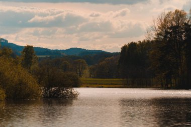 Sessiz, bulutlu bir gökyüzünü yansıtan sakin bir su kütlesi. Yumuşak ve sıcak bir ışık. Ön planda, bol miktarda zeytin yeşili çalılar kısmen suya batmış, bu da sonbaharın başlarında yeşillik ve ağaçların çeşitli karışımlarına yol açıyor.