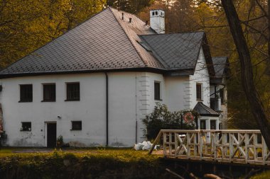 Görkemli, beyaz duvarlı, koyu kiremitli çatısı ve belirgin beyaz bacası olan bir bina, altın-kahverengi sonbahar yeşillikleri olan ağaçlardan oluşan bir zemin üzerine kurulmuş. Binanın birden fazla koyu çerçeveli penceresi ve siyah bir kapısı var.
