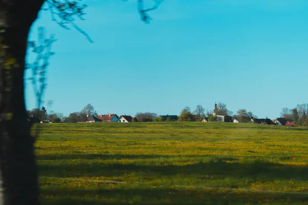 berrak, parlak mavi gökyüzünün altında sakin bir kırsal manzara. Uçsuz bucaksız yeşil bir alan, küçük sarı çiçeklerle benekli, ön plana hükmediyor, ince dalgalı desenler gösteriyor. Orta mesafede, büyüleyici bir köy ufuk boyunca uzanır, V 'nin yer aldığı.