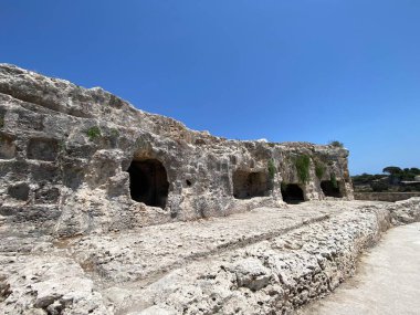 Sicilya, Syracuse 'da, açık mavi gökyüzünün altında latomie del paradiso' nun birden fazla oyulmuş açılışını gösteren yıpranmış kaya yüzeyinin alçak açılı görüntüsü.