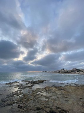 Santa Croce Camerina, Sicilya, İtalya 'daki kayalık bir plajda dramatik bulutlar arasında parlayan güneş ışınları.