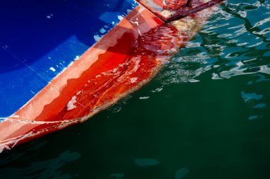 Sudan çıkan renkli tekne gövdesinin yakın plan görüntüsü yansıtıcı yüzeyleri ve deniz yüzeyinde oynayan dramatik ışığı vurguluyor.