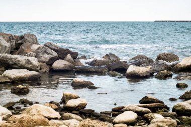 Yumuşak dalgalı puslu deniz manzarası kayalık kıyı şeridini okşayarak bulutlu gökyüzü altında sakin kıyı atmosferini somutlaştırıyor.