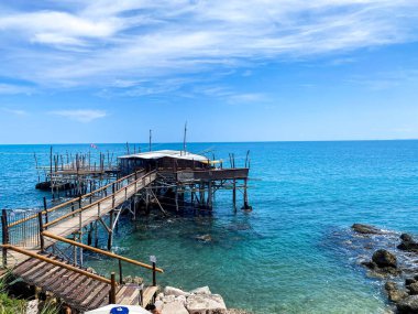 Turkuaz sulara uzanan ahşap iskele, Rocca San Giovanni kıyı manzarasında mavi gökyüzüne karşı geleneksel avlanma platformu Trabocco