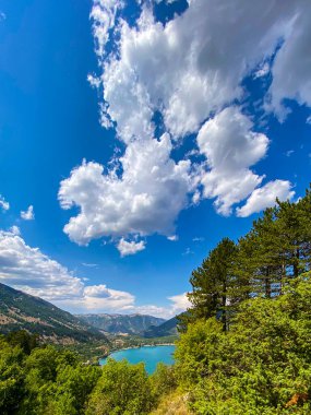 Scanno Gölü 'nün nefes kesen manzarası, bereketli bitki örtüsüyle çevrili ve üzerinde beyaz bulutlar olan canlı mavi bir gökyüzü ile çevrili.