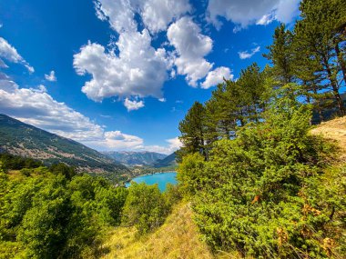 Çam ağaçları, İtalya 'nın Abruzzo kentindeki dağlık arazide, parlak mavi gökyüzünün altında kurulmuş Scanno Gölü' nün nefes kesici manzarasını resmediyor.