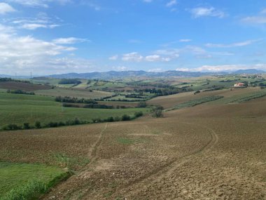 Yuvarlanan tepeler, tarlaları ve yeşil tarlaları gösteriyor. Parlak bir gökyüzünün altında, kırsal bölgelerde yumuşak bulutlar var.