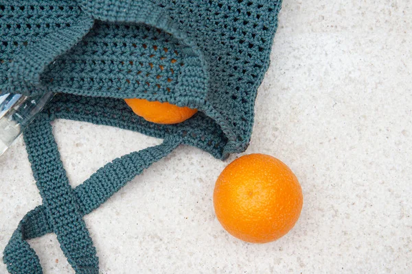 Grey knitted bag handmade, ripe oranges and a bottle of water outdoors. Sustainable shopping. Waste-fr ee lifestyle. Do-it-yourself jute bag