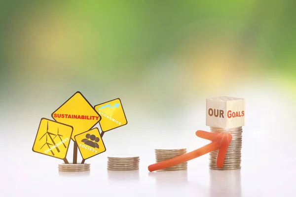 Our goals on cube on stack of coins with environment, society and economics written on yellow sign and arrow upwards on abstract background. Sustainable with return on investment concept and economic growth idea