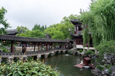 Şangay, Çin - Haziran 06, 2024: Ahşap köprüsü ve arka planda geleneksel Çin binası olan huzurlu bir gölet. Bereketli yeşillik ve canlı çiçekler huzur ortamını çevreledi..