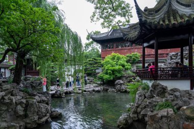 Şangay, Çin - Haziran 06, 2024: Arka planda geleneksel bir Çin pavyonu bulunan, kaya oluşumları ve yemyeşil yemyeşil ile çevrili sakin bir gölet. Ziyaretçiler Şangay 'daki Yu Garden' da geziyorlar..