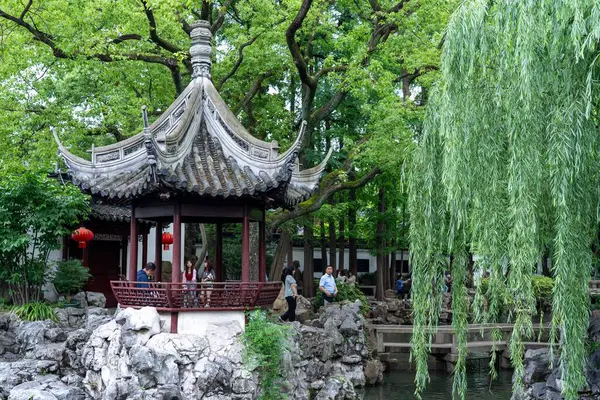 Stock image Shanghai, China - June 06, 2024 : A traditional Chinese pavilion with a curved roof stands in a lush garden with a pond, rocks and weeping willow trees.