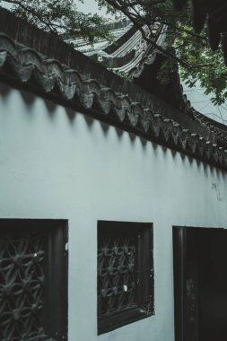 Aşağıdan iki oymalı pencere ve siyah çatı kiremitleri olan beyaz bir duvar görülüyor. Çatı yeşillikle kaplı..