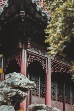 Düşük açılı kırmızı ahşap bir yapının, yaprakların arasına yerleştirilmiş, karmaşık oymalar. Yapıda bir kafes penceresi var ve bir kaya ve bulanık bir ağaç dalı tarafından kısmen gizlenmiş..