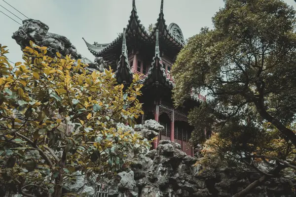 stock image A traditional Chinese pagoda with a curved roof stands nestled among lush green foliage and rocky outcroppings.