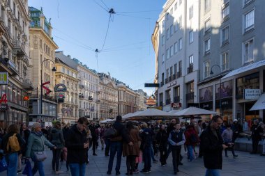 Wien, Avusturya - 28 Ekim 2023: Viyana, Avusturya 'da kalabalık bir sokak manzarası, insanların dükkanların ve kafelerin önünden geçtiği. Yüksek binaların süslemeleri var, ve gökyüzü parlak mavi.